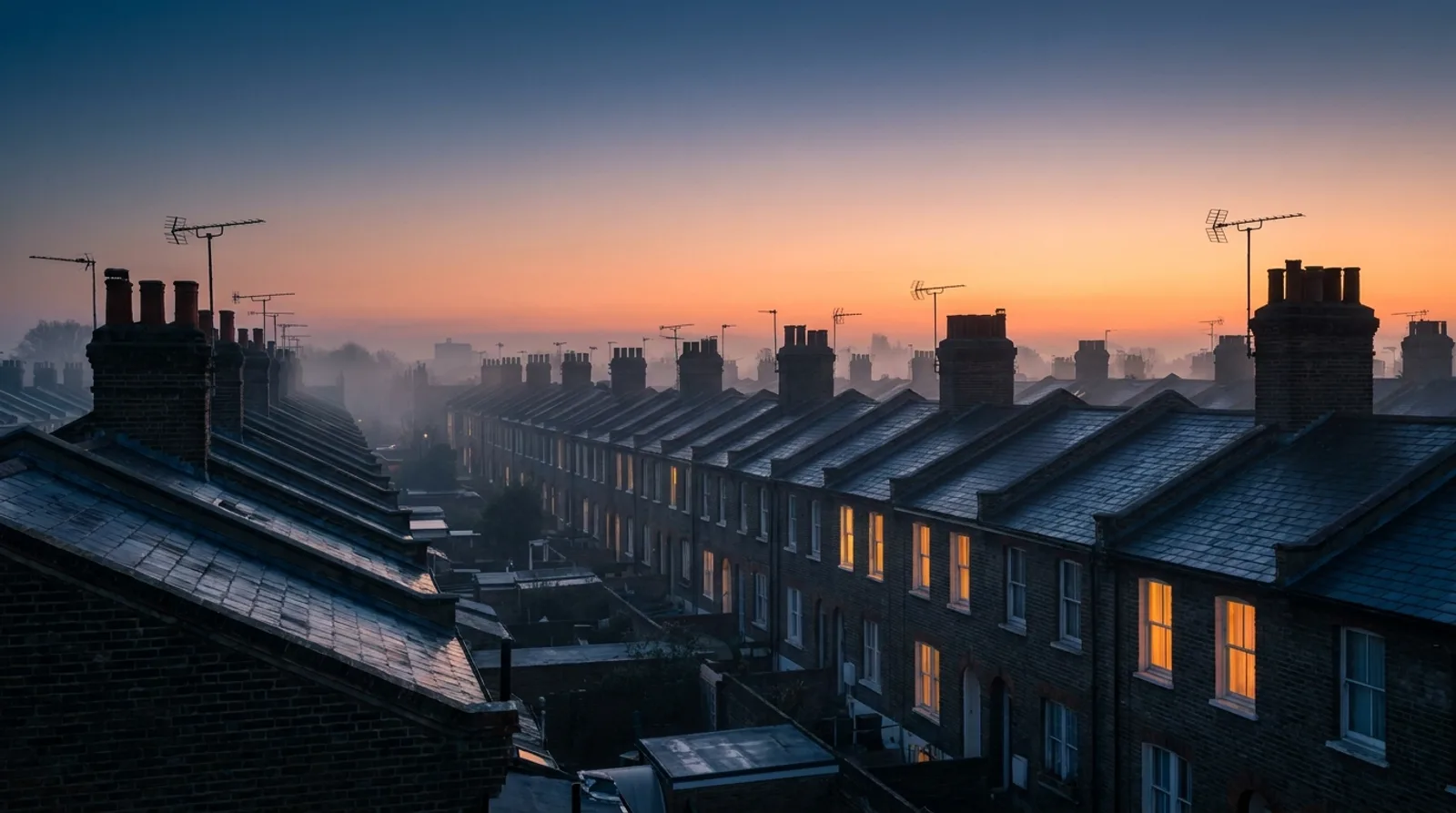 Rows of London terraced rooftops at dawn, windows glowing warm through the mist